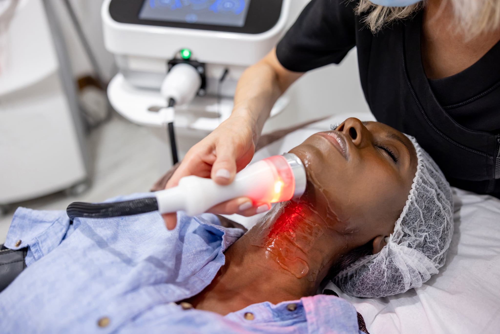 Афроамериканка в спа-салоне проходит омолаживающую процедуру на шее с помощью ультразвука и светотерапии.